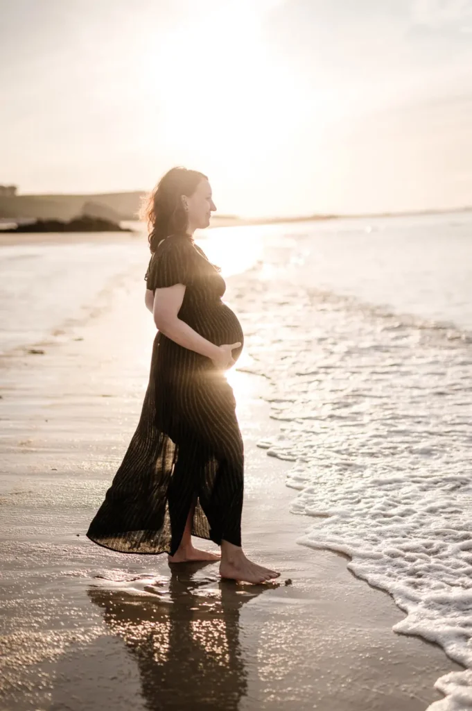 Femme enceinte qui tient son ventre les pieds dans l'eau face à la mer avec un coucher de soleil. Photos de grossesse en extérieur à Nantes